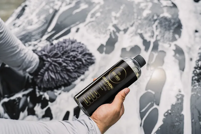 A Magic Shield Graphene Shampoo bottle held beside a vehicle during a foam wash.
