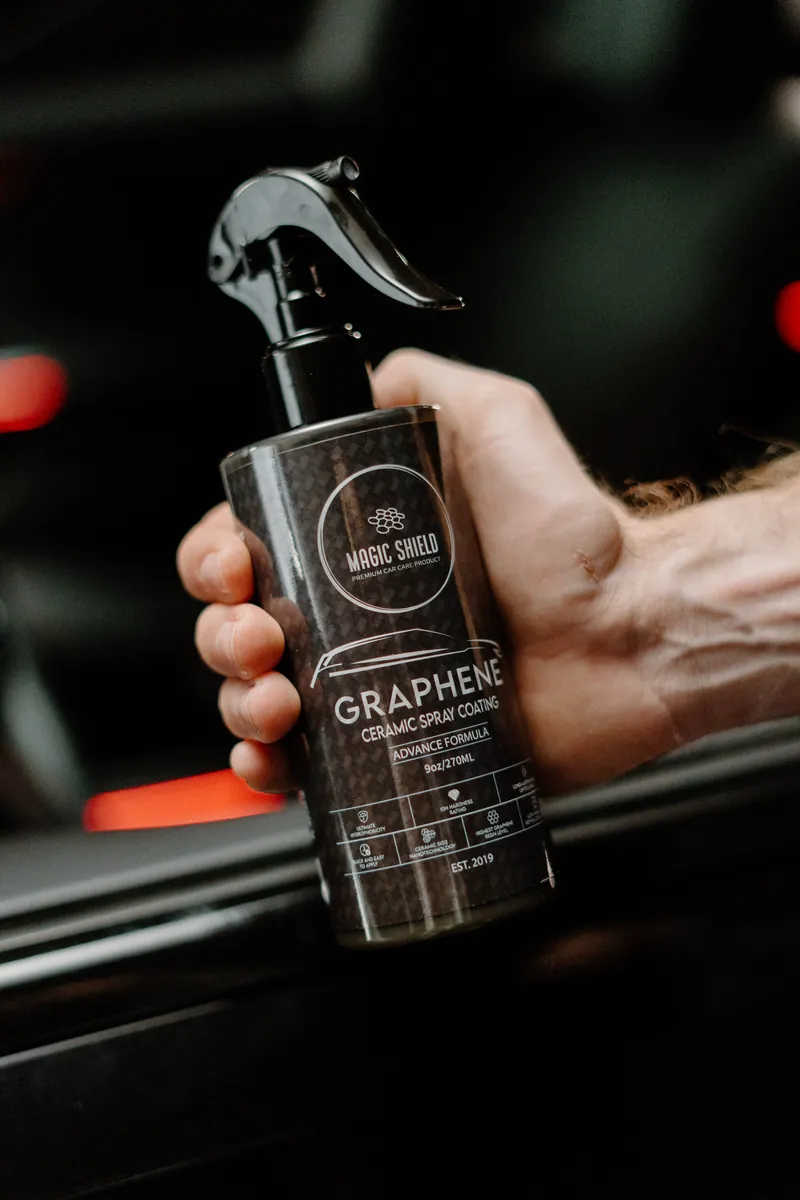 A person holding a Magic Shield Graphene Ceramic Spray bottle beside a vehicle.